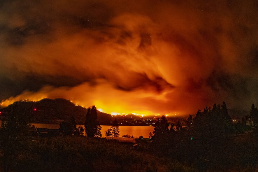 A forest fire burns among a residential area in the Okanagan Valley at night. The sky and lake glow orange.