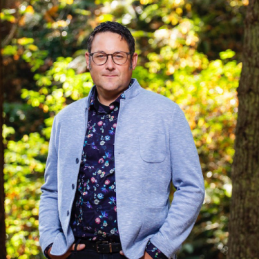 Dr. Ian Mauro stands with his hands in pockets, wearing a light blue blazer and dark blue shirt with flowers.