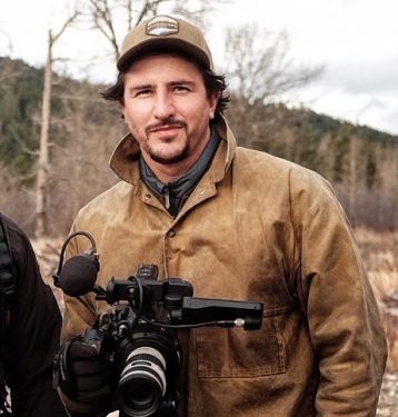 Matt Miles stands, holding a camera, wearing a tan coat and cap. Behind him there is a fall landscape in hues of brown and green.