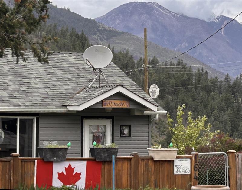 A rare surviving house in Lytton, B.C. sits behind a brown wooden fence with a metal chain-link gate. Two satellite dishes sit on the roof. A plaque above the white door reads “Miracle House.” A Canada flag hangs on the fence, with planters perched on top. Mountains with low clouds rise in the distance.