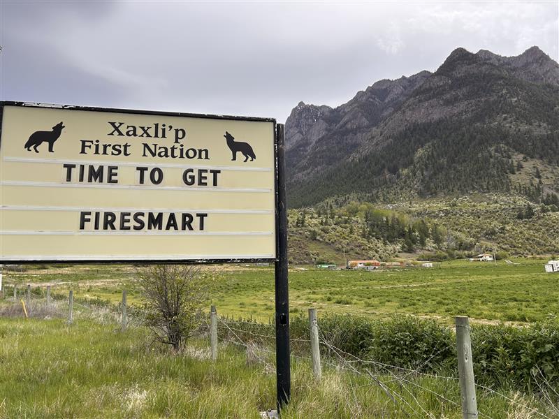 A cream-coloured sign reads “Xaxli’p First Nation – Time to Get Fire Smart,” with two howling wolf silhouettes flanking the Nation's name. Behind it lies a green field, scattered buildings, and a mountain with a rocky peak and forested base.