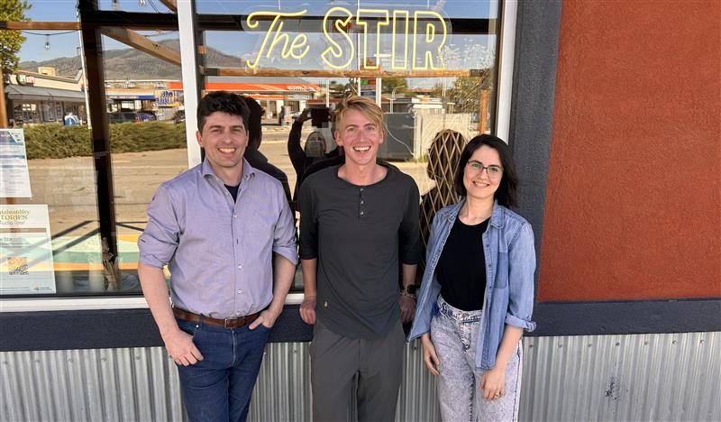 Three people stand in front of a reddish-orange building with a neon “The Stir” sign in the window. From left to right: Daniel wears jeans, a brown belt, and a grey button-up shirt, hands in his pockets. Kent Fawcett leans against the wall in dark clothes, with shaggy blonde hair. Saeideh Hejazi wears light-wash jeans, a black T-shirt, and an open denim shirt. She has a short bob and dark-rimmed glasses.