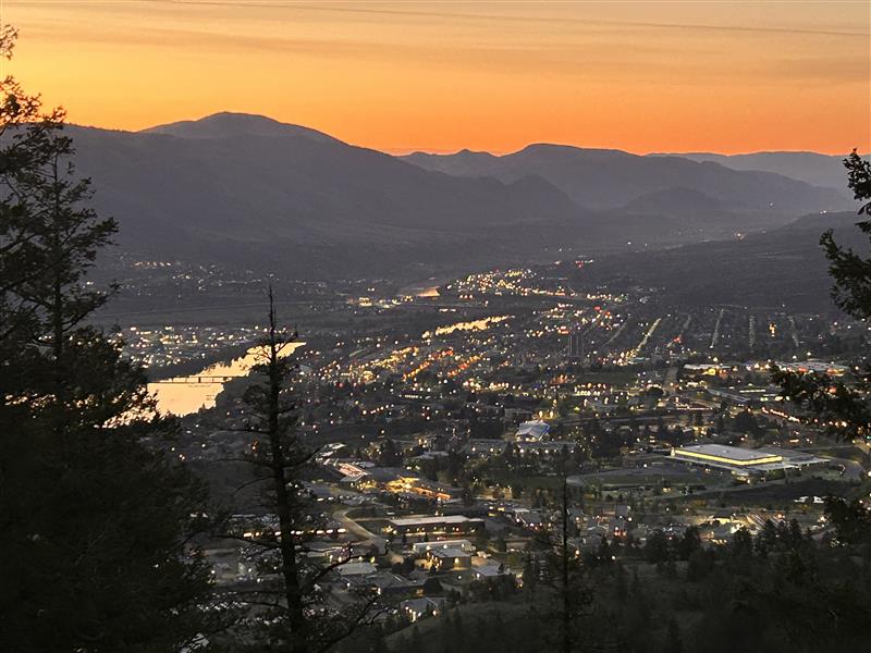 A sunrise over Kamloops casts an orange glow across the sky. City lights begin to flicker off below, with dark mountain silhouettes contrasting against the bright sky. The aerial view is from a mountain across the valley.