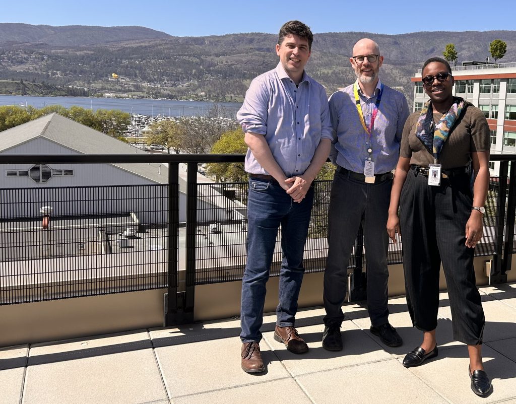 Three people stand on a rooftop in the Okanagan, smiling with the Thompson River, city buildings, and mountains behind them. Daniel is on the left, hands clasped, wearing brown dress shoes. In the middle is Julian Mallinson, bald with a grey beard, wearing glasses, a lanyard, and black sneakers. On the right, Glory Apatanku wears black dress pants, a green T-shirt tucked in, and a colorful scarf covering part of her ID lanyard. She wears sunglasses and a watch.