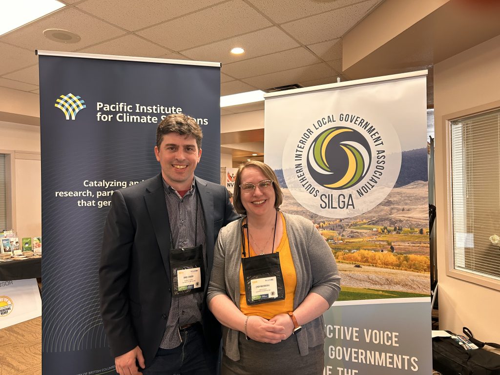 Daniel and Cindy stand in front of two banners indoors—one for the Pacific Institute for Climate Solutions, the other for the Southern Interior Local Government Association. Daniel wears a dark blue shirt with a black blazer and name tag. Cindy, shorter in height, wears a mustard-yellow top with a grey cardigan and glasses. Both smile at the camera, with a conference setting behind them.