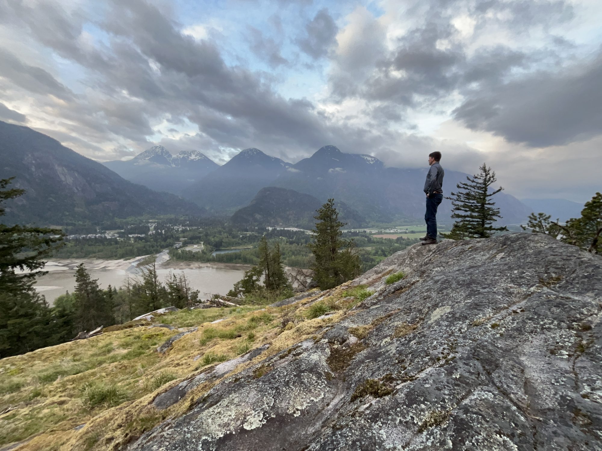 Daniel stands on the side of a rocky mountain, overlooking the Fraser River on a cloudy morning. The river winds through the valley below, with cloud-covered mountains in the distance. Daniel, seen from the side, wears jeans and a blue button-up shirt. His short brown hair is visible, but his face is turned away. His hands rest on his hips.