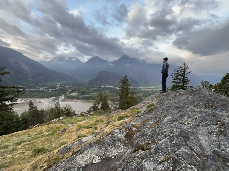 Daniel stands on the side of a rocky mountain, overlooking the Fraser River on a cloudy morning. The river winds through the valley below, with cloud-covered mountains in the distance. Daniel, seen from the side, wears jeans and a blue button-up shirt. His short brown hair is visible, but his face is turned away. His hands rest on his hips.