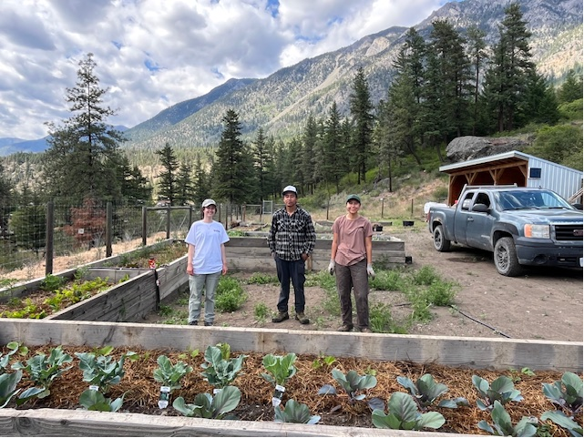 Three people stand in a semicircle of raised garden beds with visible plant growth. A navy blue pickup is parked nearby, with a white barn and mountains in the background. On the left is PICS intern Taylor Grittins in a white T-shirt, jeans, and a white baseball hat. The middle person wears a flannel shirt, jeans, boots, and a white baseball hat. On the right, a woman in gardening gloves wears dark pants, a beige T-shirt, and a green baseball cap.