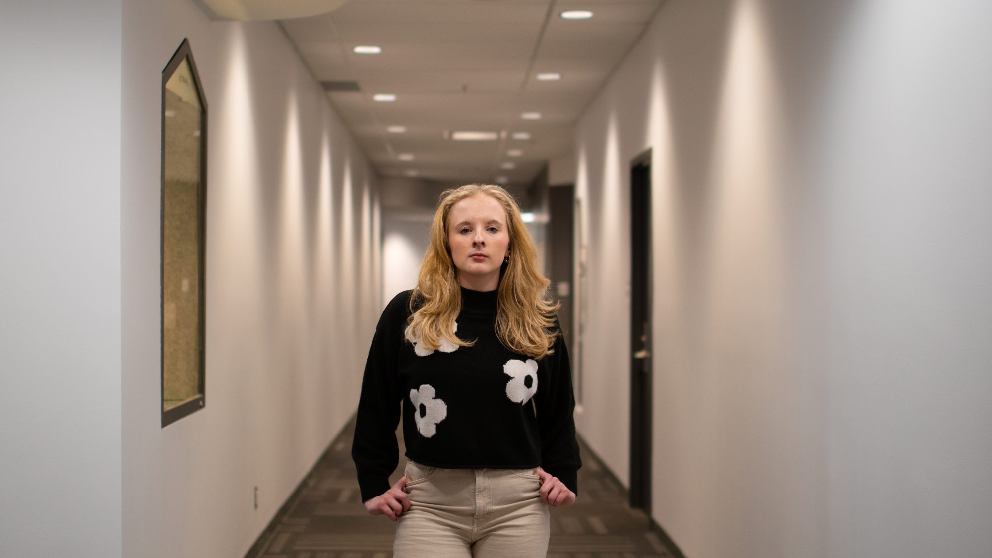 Emma Cosman stands in a white hallway with her hands by her side and a serious expression on her face. She is wearing a black sweater with white flowers and khaki pants. Her blonde hair is worn down, lying passed her shoulders.