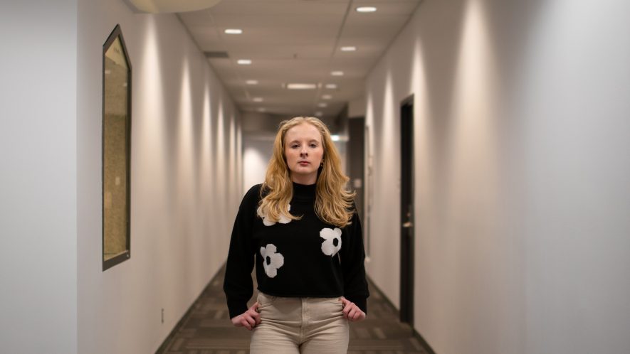 Emma Cosman stands in a white hallway with her hands by her side and a serious expression on her face. She is wearing a black sweater with white flowers and khaki pants. Her blonde hair is worn down, lying passed her shoulders.