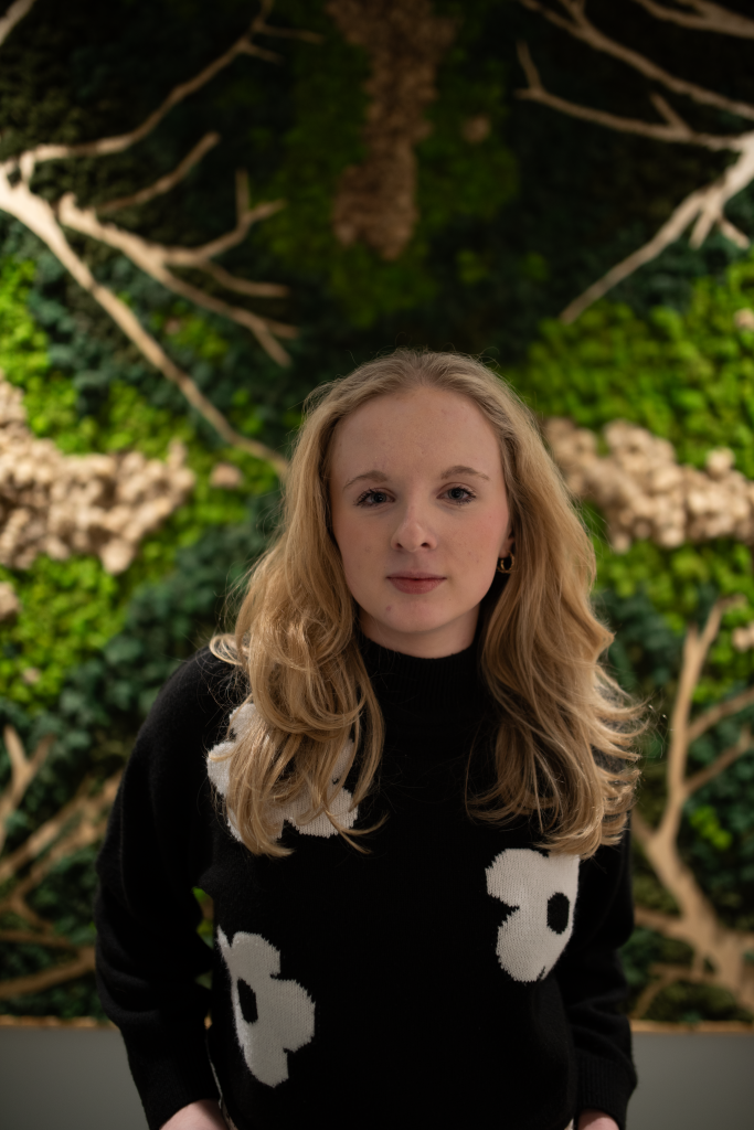 Emma stands in front of some greenery with her hands in her pockets. She is wearing a black sweater with white flowers on it. Her blonde hair is worn down past her shoulders.