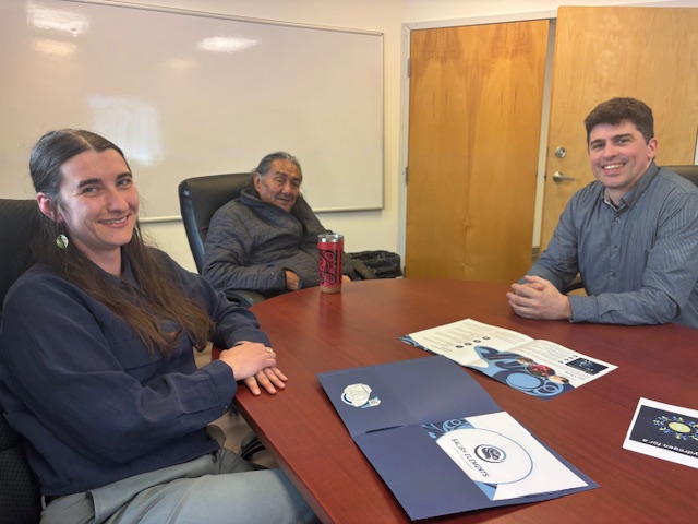 Deanna Ambrose, Xaxli’p Chief Darrell Bob Sr., and Daniel Arbour sit at a conference table smiling at the camera. Deanna wears a blue sweater, white circular earrings, and her long brown hair half-up. Chief Bob wears a grey zip-up jacket, hands in his pockets, with a red travel mug in front of him. Daniel wears a blue button-up shirt, with clasped hands. A whiteboard is behind them, with papers and infographics on the table.