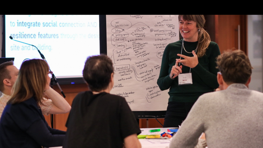 Facilitator stands smiling beside a flip chart covered with handwritten notes, leading a small group discussion at a workshop. Four participants sit around a table with markers and papers, engaged in the session."