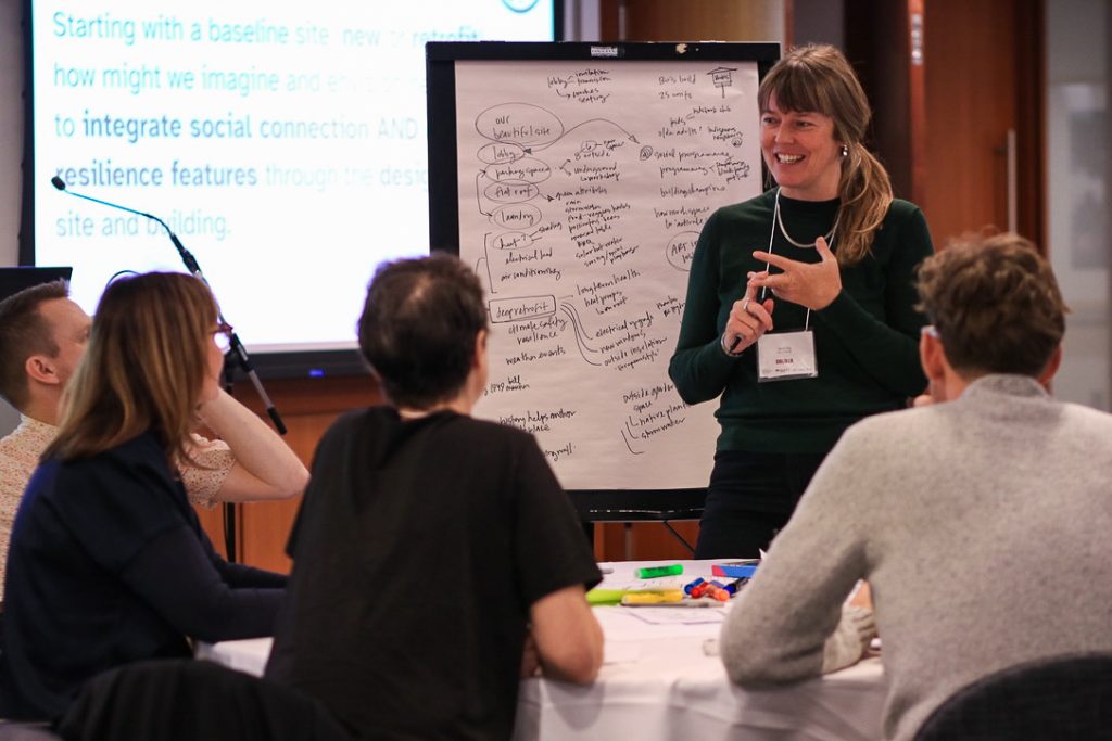Facilitator stands smiling beside a flip chart covered with handwritten notes, leading a small group discussion at a workshop. Four participants sit around a table with markers and papers, engaged in the session.