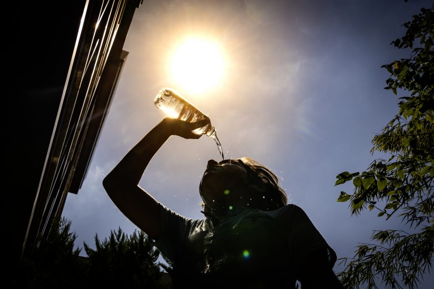 Image of a boy pouring water on his head on a hot day. The image is taken from below with the sun in the background, making the boy look like a dark silhouette.