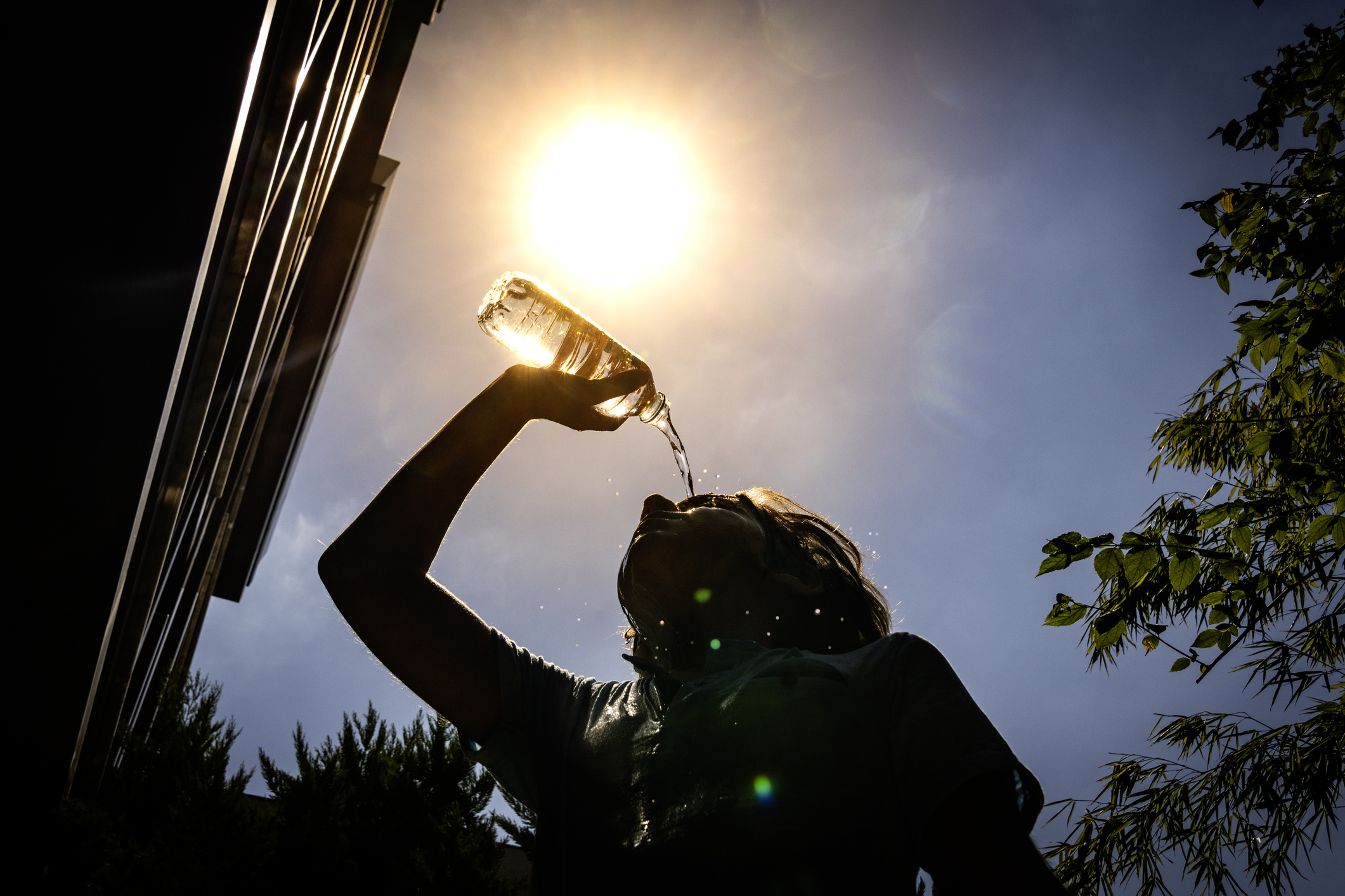 Image of a boy pouring water on his head on a hot day. The image is taken from below with the sun in the background, making the boy look like a dark silhouette.