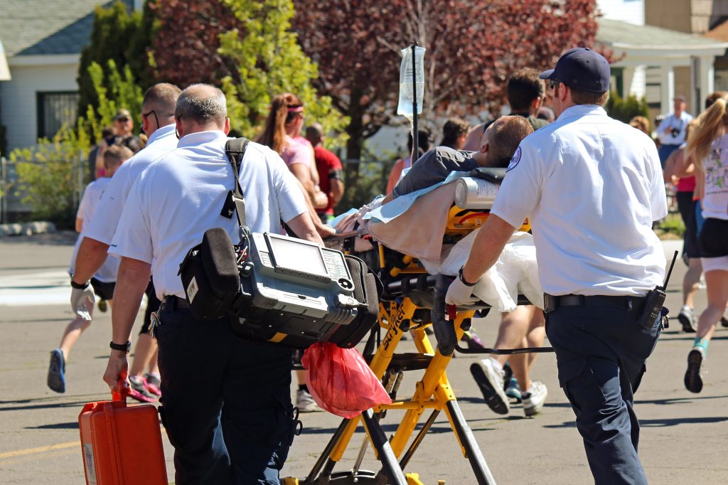 Two paramedics walk through a crowd of people with a person laying on a stretcher, there are people infront of the paramedics running in a race. It is a clear sunny day.