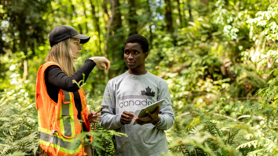 Two people standing in a lush green forest. One person, wearing an orange safety vest and black cap, gestures while speaking. The other, holding a tablet and wearing a grey T-shirt with a maple leaf and the word “Canada,” listens attentively.