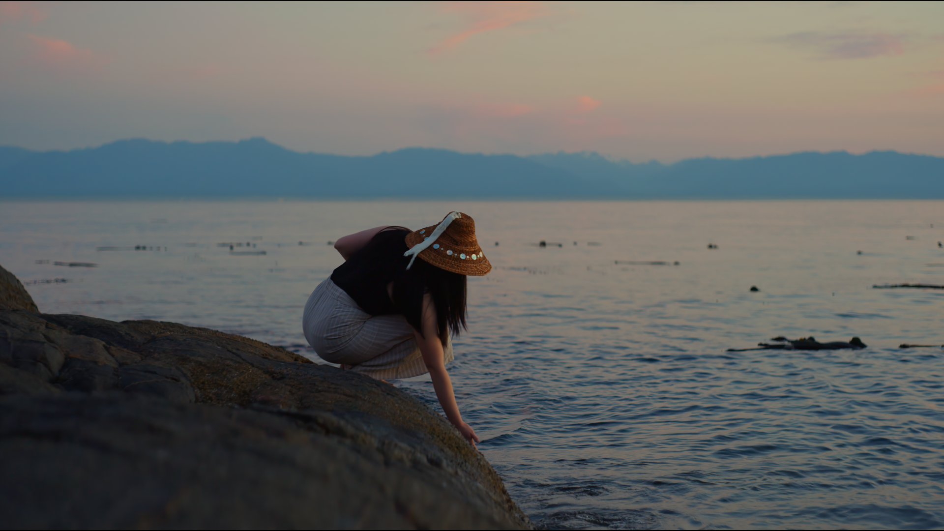 Janna Wale, wearing a cedar hat and casual clothes, leans down to touch the ocean, while crouching on a rock on the shore. Behind her, sunset fades to twilight.