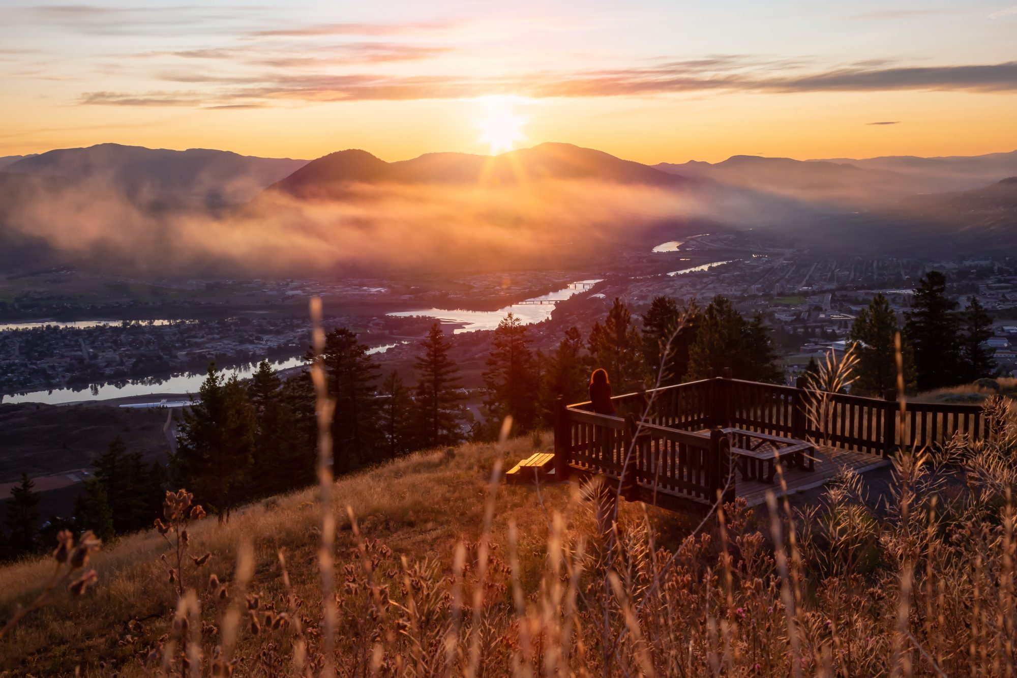 Sunset over the Okanagan Valley with a woman standing at a lookout point, viewed from behind.