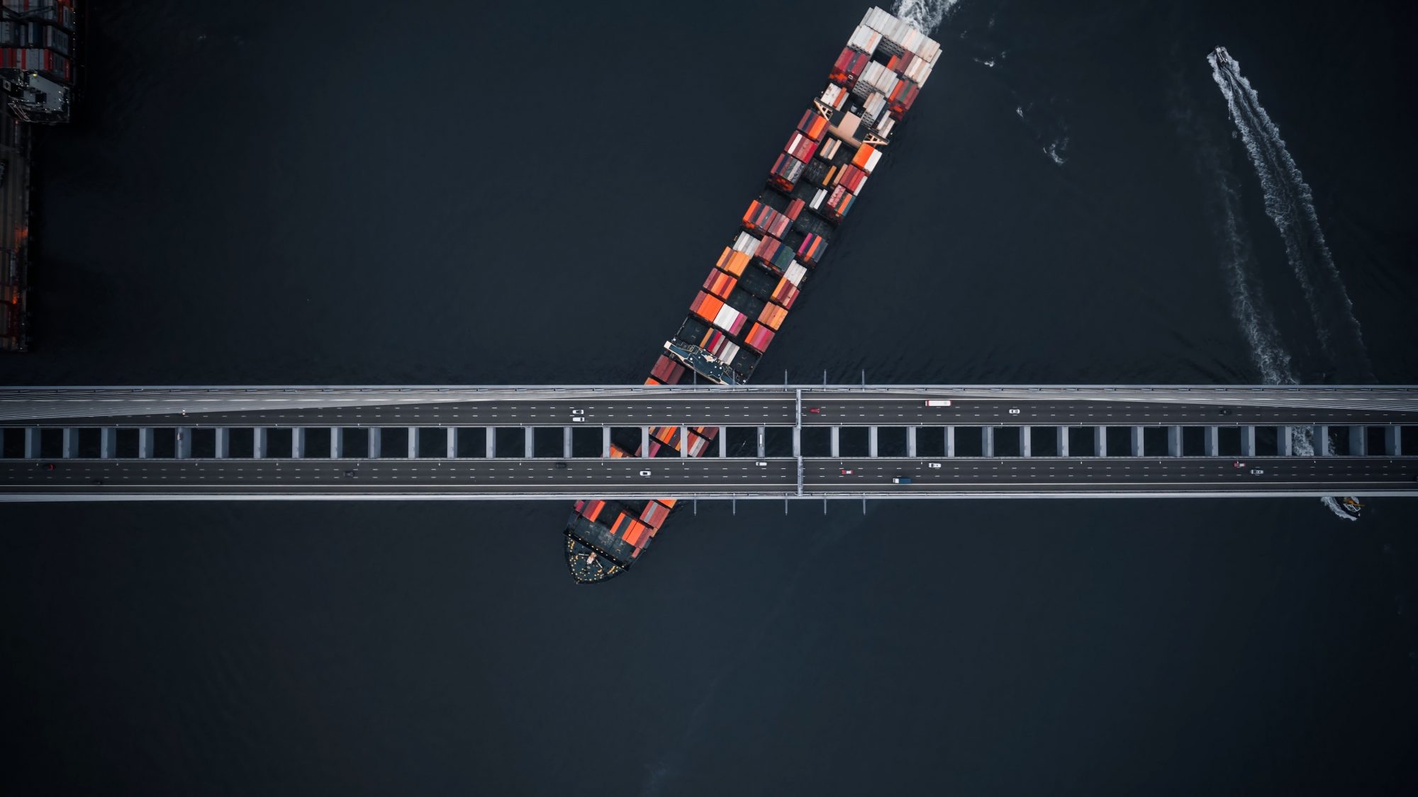 A shipping vessels sails beneath a commuter bridge with cars zipping across.