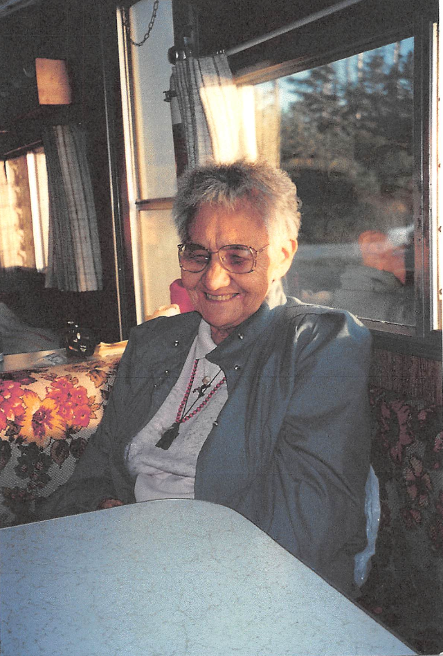 Dorothy Bell sits in a chair by a window with a table in front of her. She is smiling and looking down. She has glasses, short white hair, a white shirt, a grey jacket and multiple beaded necklaces. 