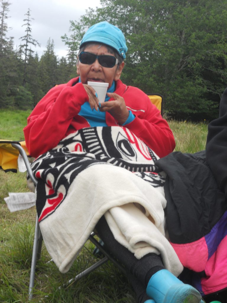 Photo of Mavis Bell sitting in a lawn chair, sipping a drink from a cup. She is wearing a blue hat, a blue shirt, a red coat, and blue rubber boots. She has a blanket across her lap and sunglasses on her face. 